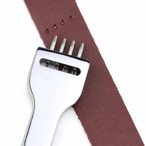 May include: A silver leather punch tool with five prongs, used for making holes in leather. The tool is placed on a piece of brown leather.