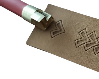 May include: A close-up of a leather stamping tool being used to create a decorative pattern on a piece of brown leather. The tool has a wooden handle and a metal head with a geometric design.