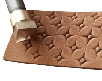 May include: A close-up of a leather stamping tool with a brass head and a wooden handle. The tool is being used to create a pattern of small circles and four-leaf clover shapes on a piece of brown leather.