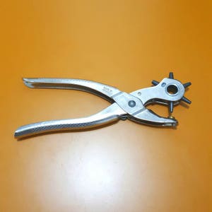 May include: A silver-colored leather punch tool with multiple hole size options. The tool has long handles for leverage and a rotating head with various sized punches. The tool is set against an orange background.