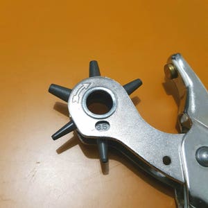 May include: A silver metal rotary punch tool with a black rotating head and a handle. The head has six black posts and a central hole. The size is marked as 3.5.