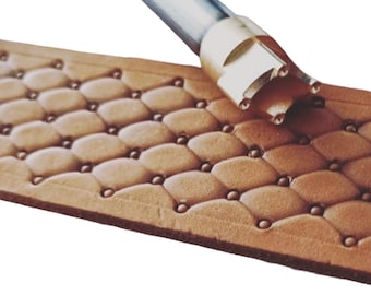 May include: A close-up of a leather stamping tool with a metal handle. The tool is used to create a diamond pattern on leather. The tool is shown on a piece of brown leather with a diamond pattern.