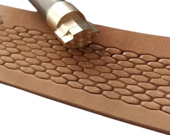 May include: A close-up of a leather stamping tool with a hexagonal pattern. The tool is made of metal and has a wooden handle. The tool is being used to stamp a piece of leather.