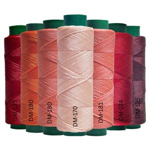 Op de afbeelding: Een verzameling kleurrijke garenklosjes in tinten rood, oranje en roze. Elke klos heeft een groene basis en is gelabeld met een DM-nummer. De draad is strak gewikkeld, waardoor een textuur ontstaat.