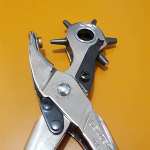May include: A silver and black Selzer punch tool with a rotating head and multiple punch sizes. The tool has a handle and is marked "Made in Germany Since 1823". The tool is designed for making holes in leather, fabric, or other materials.
