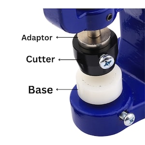 Peut inclure: Gros plan sur une machine à boutons en métal bleu avec une base blanche, un coupe-bouton noir et un adaptateur argenté. La machine à boutons est utilisée pour créer des boutons personnalisés.