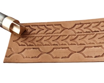 May include: A close-up of a leather stamping tool being used to create a decorative pattern on a piece of brown leather. The tool has a brass handle and a metal head with a geometric design.