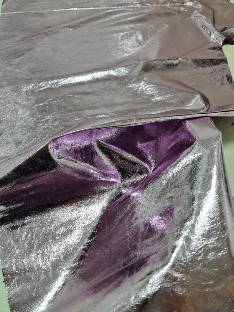 May include: A piece of silver metallic leather with a purple hue. The leather is folded and wrinkled, showing its texture.