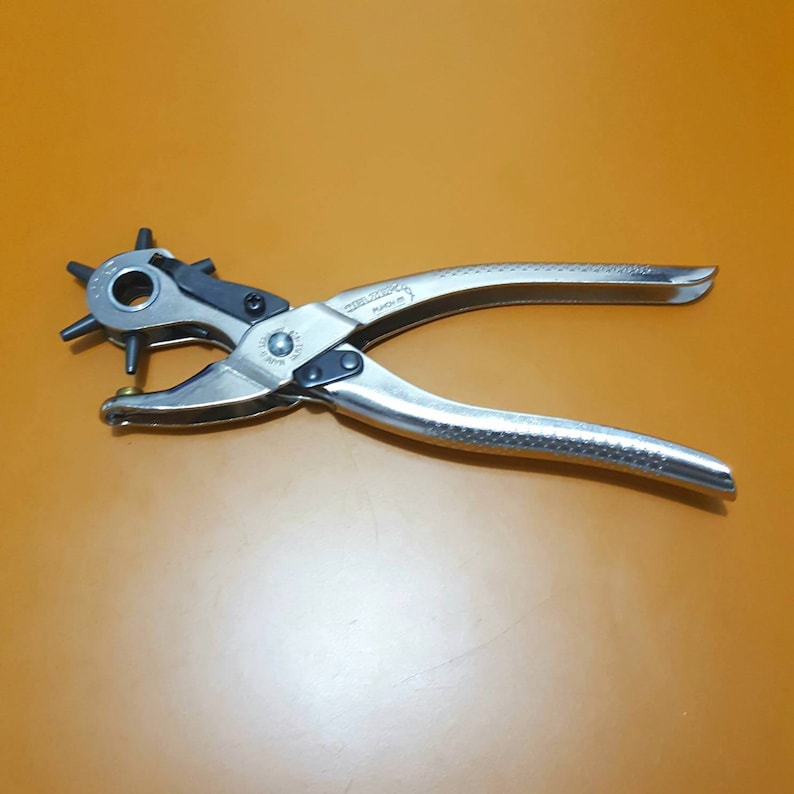 May include: A silver-colored multi-size hole punch tool with black accents. The tool has a rotating head with multiple punch sizes and textured handles for grip. The tool is designed for punching holes in leather, paper, or other materials.