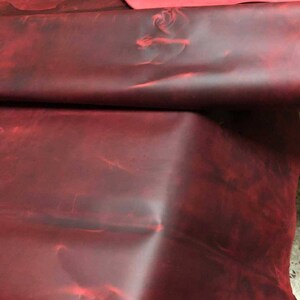 May include: A close-up of a deep red leather hide with a mottled, marbled appearance. The leather is folded and draped, showcasing its texture and color.