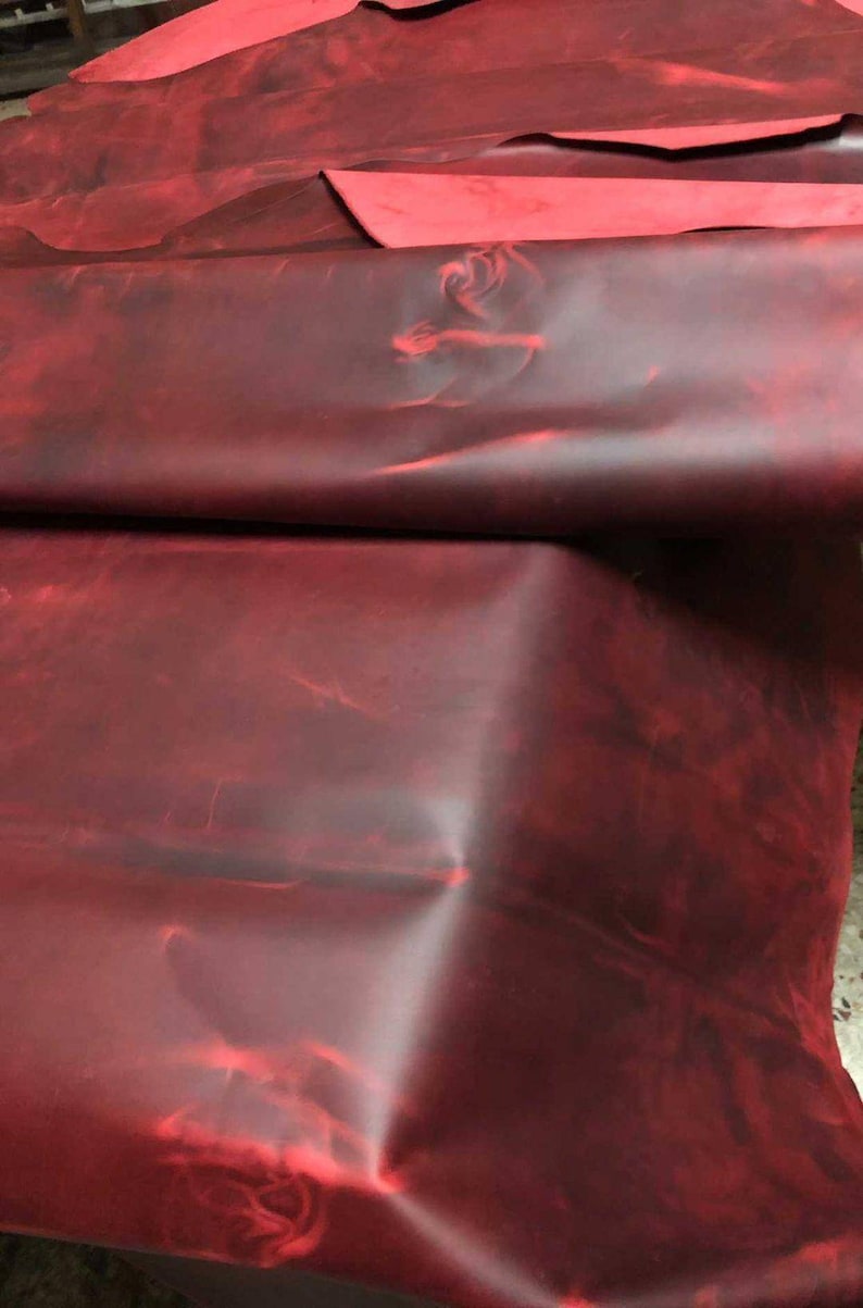 May include: A close-up of a deep red leather hide with a mottled, marbled appearance. The leather is folded and draped, showcasing its texture and color.