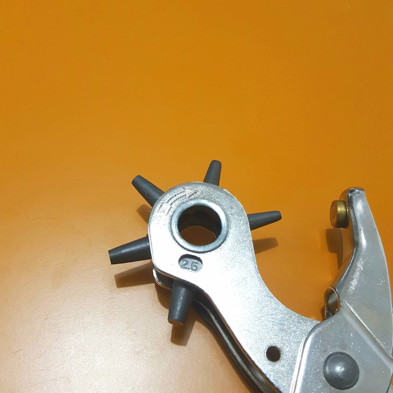 May include: A silver metal hole punch tool with a rotating head featuring multiple hole sizes. The tool has a circular punch head with six black rubber tips. The number "25" is visible on the tool. The background is a solid orange color.