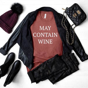 May include: A rust-colored t-shirt with the text "MAY CONTAIN WINE" in white lettering. The shirt is styled with a black leather jacket, black jeans, a black purse with a gold chain strap, and a burgundy beanie with a black pom-pom.