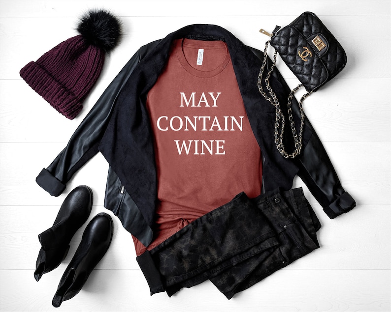 May include: A rust-colored t-shirt with the text "MAY CONTAIN WINE" in white lettering. The shirt is styled with a black leather jacket, black jeans, a black purse with a gold chain strap, and a burgundy beanie with a black pom-pom.