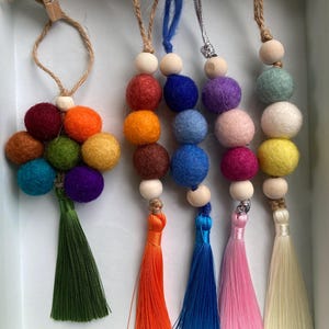 May include: A selection of colourful felt ball garlands with wooden beads and tassels. The garlands feature a variety of colours, including orange, blue, pink, and green. The garlands are displayed in a white box.