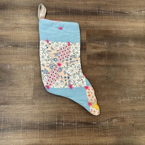 May include: A quilted Christmas stocking featuring a light blue top cuff and a patchwork body composed of various floral and geometric patterned fabrics in shades of white, red, and yellow, accented with small pink yarn ties at the quilt intersections.