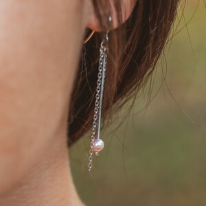 Puede incluir: Un pendiente de cadena de plata con una pequeña perla colgando del extremo. El pendiente se lleva en la oreja izquierda de una persona con cabello castaño.