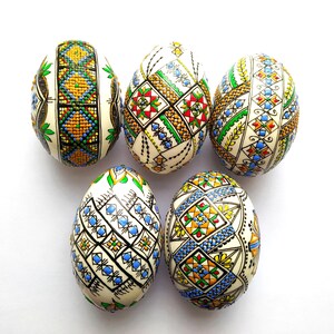 May include: Five intricately decorated Easter eggs with colorful geometric patterns. The eggs feature designs in shades of blue, green, yellow, red, and black, set against a cream-colored background. The eggs are arranged on a white surface.