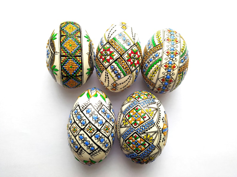 May include: Five intricately decorated Easter eggs with colorful geometric patterns. The eggs feature designs in shades of blue, green, yellow, red, and black, set against a cream-colored background. The eggs are arranged on a white surface.