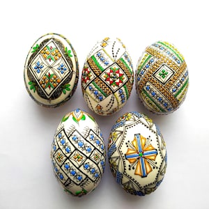 May include: Five decorated Easter eggs with intricate patterns in various colors, including blue, green, red, and gold. The eggs have a cream-colored base and are arranged on a white surface. The designs feature geometric and floral motifs.