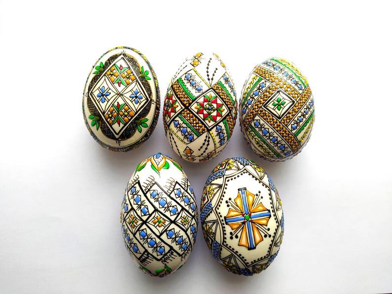 May include: Five decorated Easter eggs with intricate patterns in various colors, including blue, green, red, and gold. The eggs have a cream-colored base and are arranged on a white surface. The designs feature geometric and floral motifs.