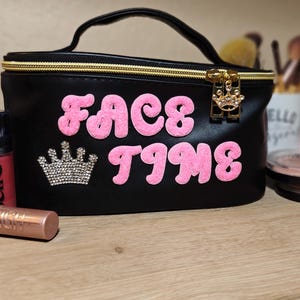 Peut inclure: Trousse de maquillage noire avec l'inscription "FACE TIME" en lettres roses et bouffantes. Le sac est doté d'une fermeture éclair dorée et d'un petit charme en forme de couronne. Une couronne en strass est en dessous.