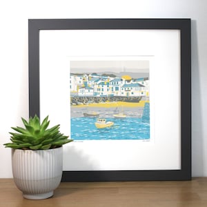 May include: A framed print of a colorful illustration of a seaside town with boats in the harbor. The print is in a black frame and has a white mat. The title of the print is "South Cornwall" and the artist's name is "Joe Wright".