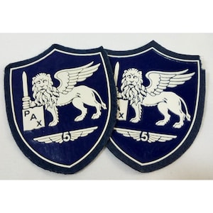 Puede incluir: Dos parches azul marino con un león alado blanco sosteniendo una espada y una pancarta con las letras "PAX". Los parches tienen forma de escudo con un borde blanco y el número "5" en la parte inferior.