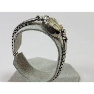 May include: A silver wristwatch with a square face and a silver metal band. The watch is on a white display stand.