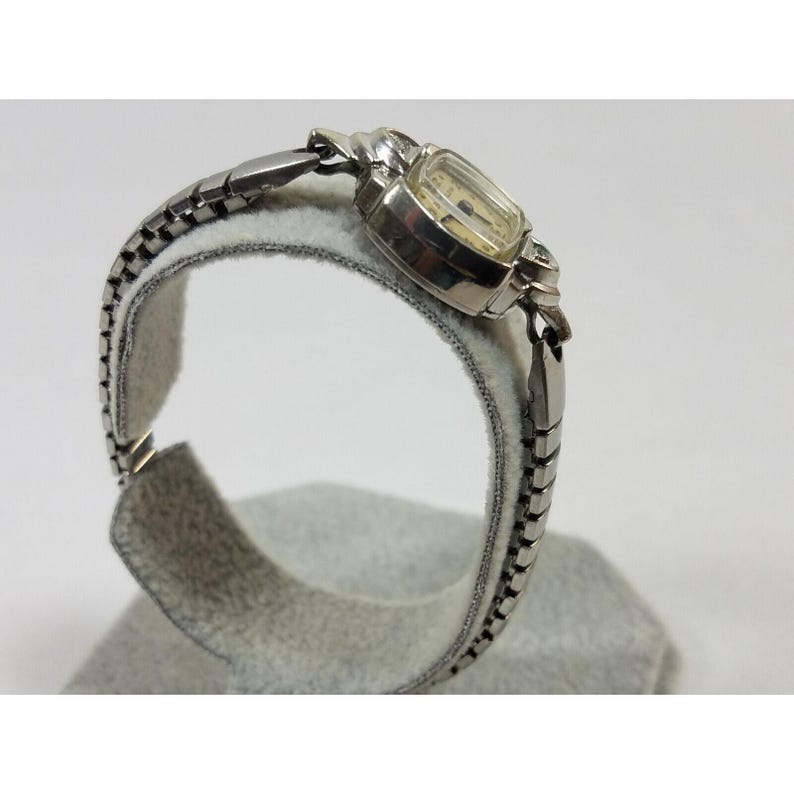 May include: A silver wristwatch with a square face and a silver metal band. The watch is on a white display stand.