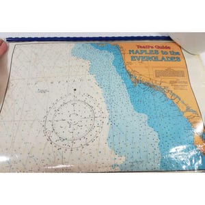 Puede incluir: Una carta náutica titulada "Teal's Guide Naples to the Everglades" con colores azul y beige. La carta presenta líneas detalladas, números y una rosa de los vientos, que indica la profundidad y la información de navegación. La carta está protegida por una lámina de plástico transparente.
