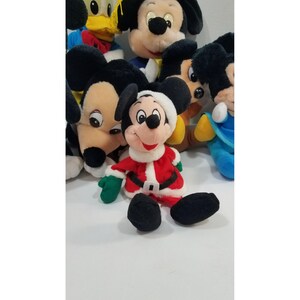 May include: A collection of plush Mickey Mouse toys. The central Mickey is dressed in a Santa Claus outfit with a red coat, black belt, and green gloves. Other Mickey Mouse plush toys are visible in the background.