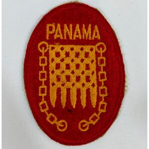 Pode incluir: Remendo vermelho oval com a palavra "PANAMA" bordada em ouro. O centro apresenta um desenho dourado com uma borda de corrente. O remendo é provavelmente um item militar ou comemorativo.