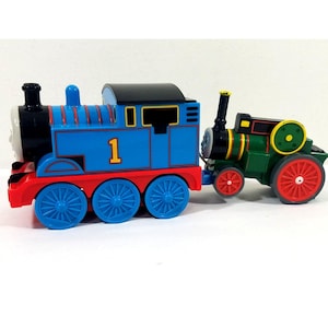 May include: A blue toy train with the number 1 on the side, red wheels, and a black top. It is connected to a green tractor with red wheels. The train has a smiling face.