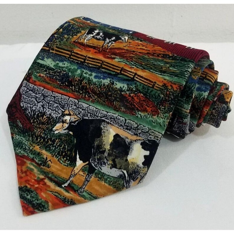 May include: A rolled-up necktie featuring a pastoral scene with cows, a wooden fence, and a field. The tie has a burgundy back and a colorful design with shades of green, orange, and brown. The tie is made of a smooth fabric.