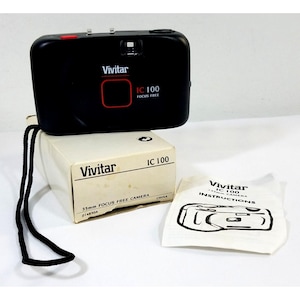 Puede incluir: Una cámara negra Vivitar IC 100 de 35 mm sin enfoque con un botón rojo, sobre su caja original y manual de instrucciones. La caja y el manual son blancos con texto negro. Se adjunta una correa de cámara negra.