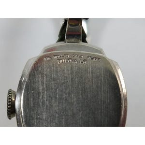 May include: A silver-toned wristwatch with a round face and a silver band. The back of the watch has the text "10k Rolled Gold Plate Bulova 16" engraved on it.
