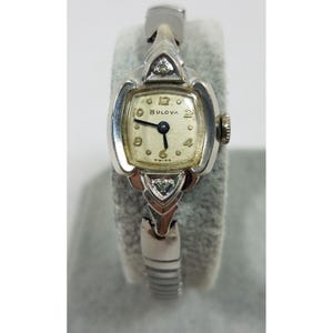 May include: A silver Bulova wristwatch with a square face and diamond accents. The watch has a white face with black Roman numeral hour markers and black hands. The watch is on a silver band.
