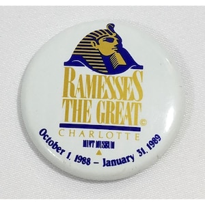 May include: White circular button featuring the text "RAMESSES THE GREAT" in gold, with a blue and gold illustration of an Egyptian pharaoh. The button also includes the text "CHARLOTTE MINT MUSEUM" and the dates "October 1, 1988 - January 31, 1989."