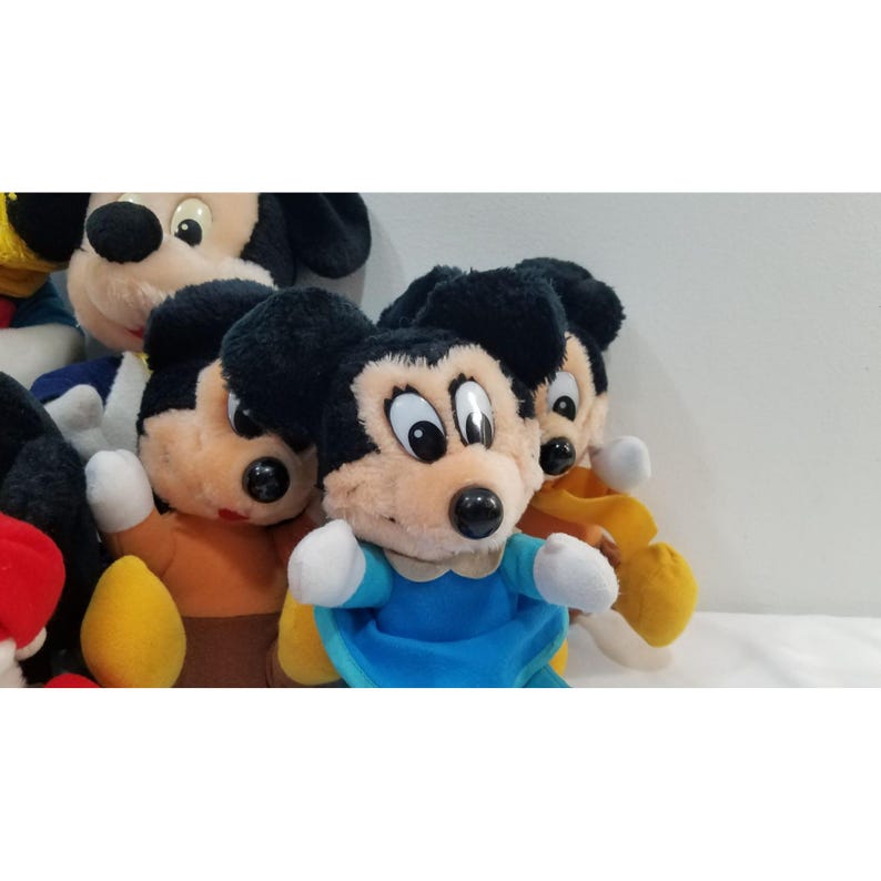 May include: A collection of Mickey Mouse plush toys. The plush toys have black ears, a tan face, and a black nose. One Mickey Mouse is wearing a blue outfit with white gloves. Other Mickey Mouse plush toys are visible in the background.