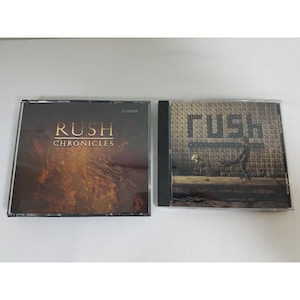 May include: Two compact discs with black cases. The left CD has a brown and gold background with the text "Rush Chronicles" in gold lettering. The right CD has a black and white image of a person walking on a tiled surface with the text "Rush" in gold lettering.