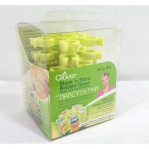 Puede incluir: Una caja de plástico transparente que contiene una torre de bobinas Clover Stack 'n Store. La torre está llena de soportes de bobinas amarillas. La caja tiene un embalaje verde y blanco con texto y una imagen de Nancy Zieman. Art. No. 9508.