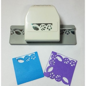 May include: A white paper punch with a gray base. The punch creates a decorative border with circles and leaves. Two samples of the punched design are shown, one on blue paper and one on purple paper.