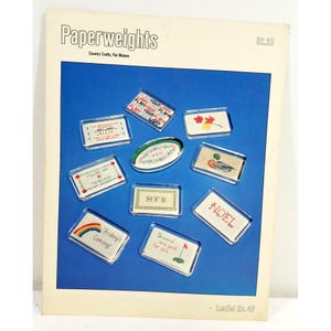 May include: A magazine cover titled "Paperweights" with various small, rectangular paperweights. The paperweights feature embroidered designs and text, including "Friday's Coming!" and "Greens are good for you." The background is a vibrant blue.