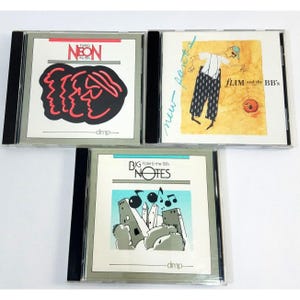 Puede incluir: Tres discos compactos con portadas de álbum que presentan ilustraciones. La primera portada de álbum es roja y negra con el texto "Flim Neon the 00s". La segunda portada de álbum es amarilla y presenta un personaje de dibujos animados con el texto "Flim and the BB's". La tercera portada de álbum es azul y gris con el texto "Big Flim & the 00s Notes".