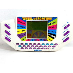 May include: A white handheld electronic game titled Wheel of Fortune by Tiger. The device features a central LCD screen surrounded by a colorful wheel graphic, a QWERTY keyboard with purple buttons, and additional purple control buttons on the sides.