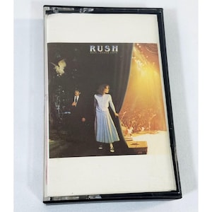 May include: A vintage cassette tape featuring the album cover for Rush's Exit...Stage Left. The cover art depicts a stage scene with figures and the band's name in a bold font. The cassette is in a black case.