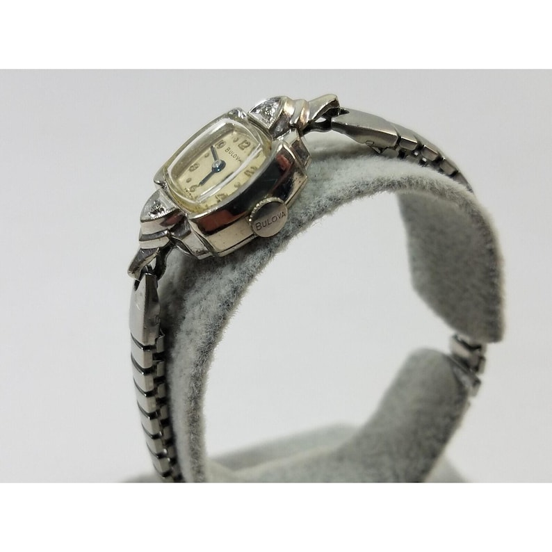May include: A silver and gold wristwatch with a square face and diamond accents. The watch face has black Roman numeral hour markers and a blue hand. The watch band is a silver metal link band. The watch is on a gray fabric surface. The watch face says "BULOVA".