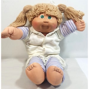 May include: A doll with light skin, green eyes, and blonde yarn hair styled in two pigtails. The doll wears a white collared shirt with purple striped sleeves and matching purple and white striped pants, featuring a soft fabric construction.