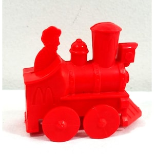 May include: A bright red plastic toy train with a silhouette of a person in the driver's seat. The train has two large wheels and the McDonald's logo on the side. The toy is a solid, vibrant red color.